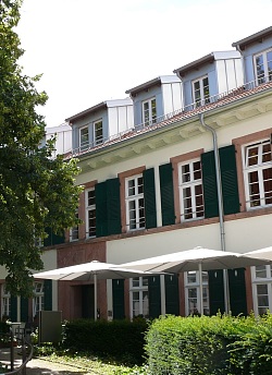 Das Bild zeigt das Alten- und Pflegeheim Spital, ein zweistöckiges gelbes Gebäude mit grünen Fensterläden und grauen Dachgauben vor dem hinter einer Hecke Sonnenschirme aufgespannt sind.