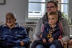 Ein älterer Mann mit weißen Haaren sitzt im Rollstuhl neben einer jüngeren Frau auf einem Stuhl. Auf ihrem Schoß ein kleiner Junge. Alle schauen in die gleiche Richtung, wo eine Aufführung oder Ähnliches stattfindet. Die beiden Erwachsenen lächeln.