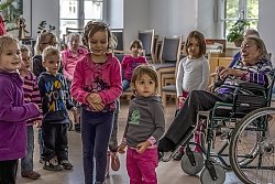 Eine Gruppe kleinerer Kinder führt ein Programm vor den Bewohnern des Spitals auf. Man sieht Jungs und Mädchen stehen und in bewegung in einem Raum. rechts eine Seniorin im Rollstuhl. Im Hintergrund weitere Kinder und Senioren.