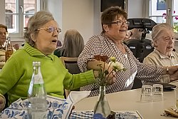 Drei Frauen sitzen an einem Tisch. Sie halten sich an den Händen, singen und schunkeln. Im Hintergrund ein weiterer Tisch, an dem ältere menschen zusammensitzen.