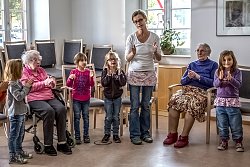 Vier kleine Mädchen stehen im Halbkreis mit ihrer Gruppenleiterin, dazwischen zwei Seniorinnen, eine im Rollstuhl. Alle klatschen und singen gemeinsam.