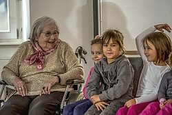 Eine Seniorin im Rollstuhl sitzt neben drei kleinen Mädchen, eine erzählt etwas und hebt den Arm über den Kopf. Die Seniorin schaut die Mädchen lachend an. Die Kinder schmunzeln.