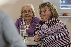 Zwei Frauen, eine ältere und eine jüngere sitzen an einem Tisch. Sie schauen beide interessiert in eine Richtung, wo eine Aufführung stattfindet.